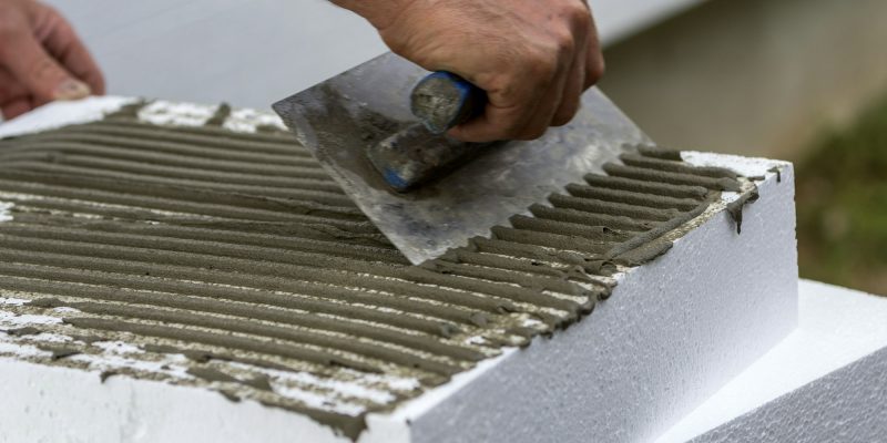 Close-up of worker hand with trowel applying glue on white rigid polyurethane foam sheet for house