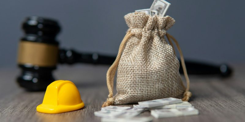 Closeup image a bag of money, hard hat and gavel on a wooden table.