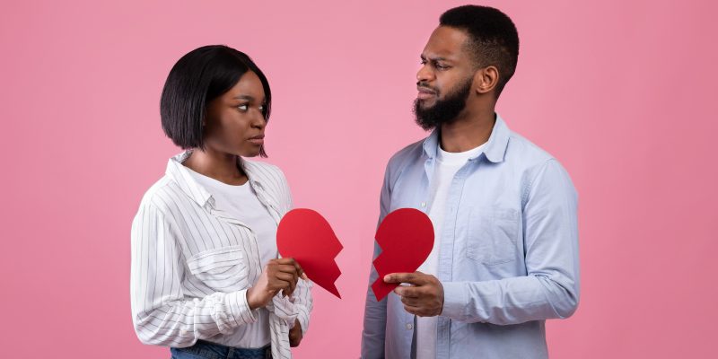 Displeased black guy and lady holding halves of torn paper heart on pink studio background. Divorce