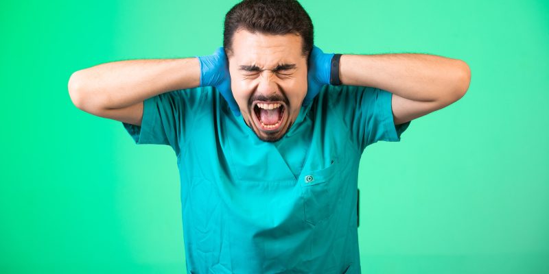 Doctor in uniform and hand mask covering his ears and having emotional shock, on green background